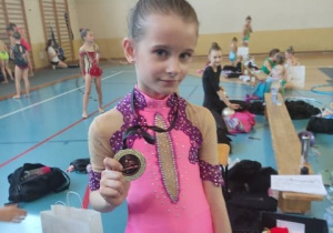 Hala sportowa, uśmiechnięta dziewczynka z dumą prezentuje swój medal.